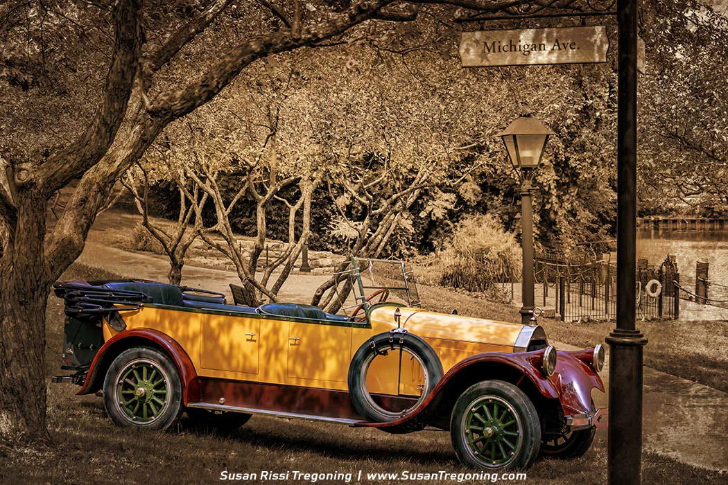 A yellow and red Pierce‑Arrow Model 33 touring car with green spoked wheels is parked on a grassy area beside a paved path in a park setting. The background is rendered in sepia while the car remains in full color, creating a selective‑color effect. Trees with full foliage, a lamppost, and a “Michigan Ave.” street sign appear behind the car, with a fence and water visible in the distance.