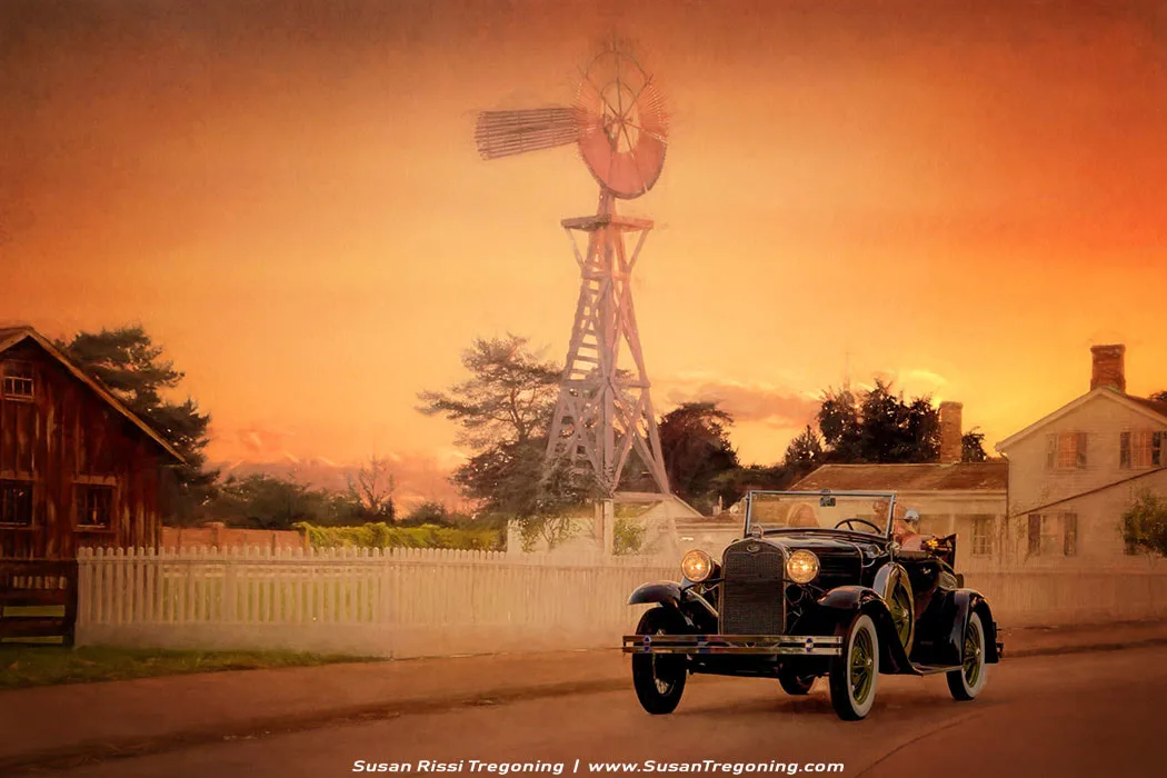 A Ford Model A Sport Coupe rolls past a white picket fence in a warm, sunset‑lit rural scene. The black convertible‑style body, upright windshield, and tall narrow wheels stand out against the soft orange and pink sky. A tall windmill rises behind the car, with a weathered wooden barn and a white farmhouse completing the early‑20th‑century countryside setting. The moment feels nostalgic and calm, as if capturing a quiet drive at the end of the day.