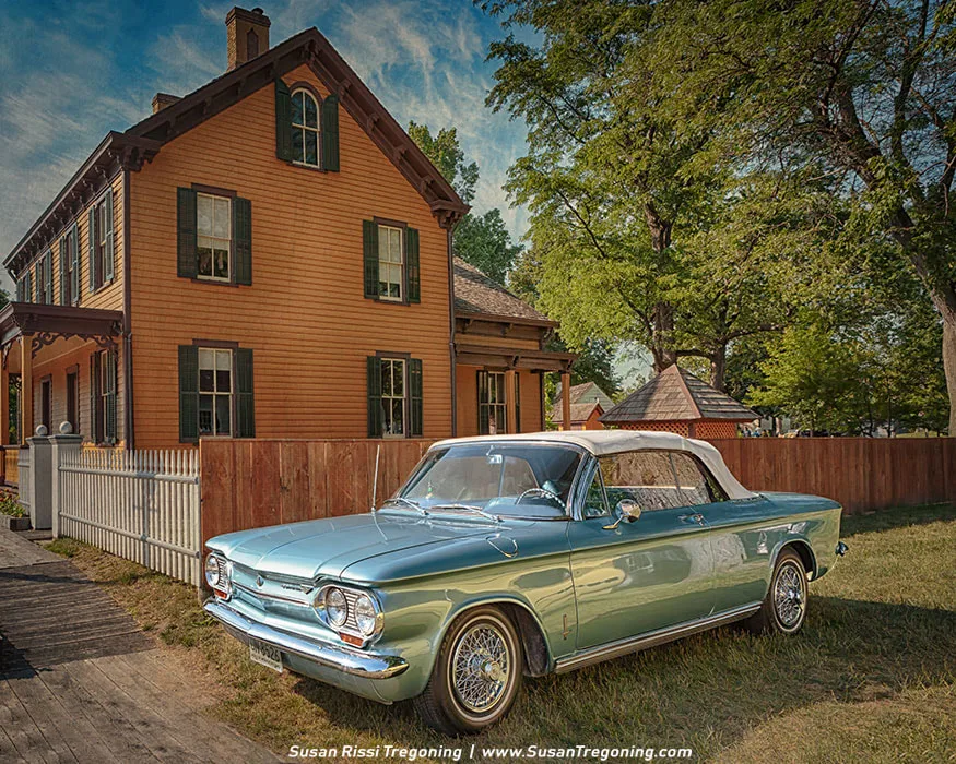 A 1963 Chevrolet Corvair convertible in light blue with a white soft top is parked on a grassy lawn in front of an orange historic house with green shutters. Chrome trim, wire‑spoke wheels, and the car’s low, streamlined profile highlight the Corvair’s early‑1960s styling. A white picket gate and wooden fence frame the house, with tall trees and additional buildings in the background, creating a nostalgic small‑town setting.