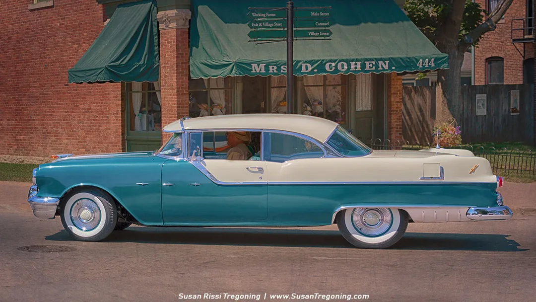 A turquoise and white 1950s-era classic car with chrome trim and whitewall tires is parked along a brick storefront marked “Mrs. D. Cohen” under green awnings. A nearby signpost points toward village attractions including a working farm, Main Street, a carousel, the exit and store, and the village green. The nostalgic small‑town setting suggests a historic or interpretive village. 