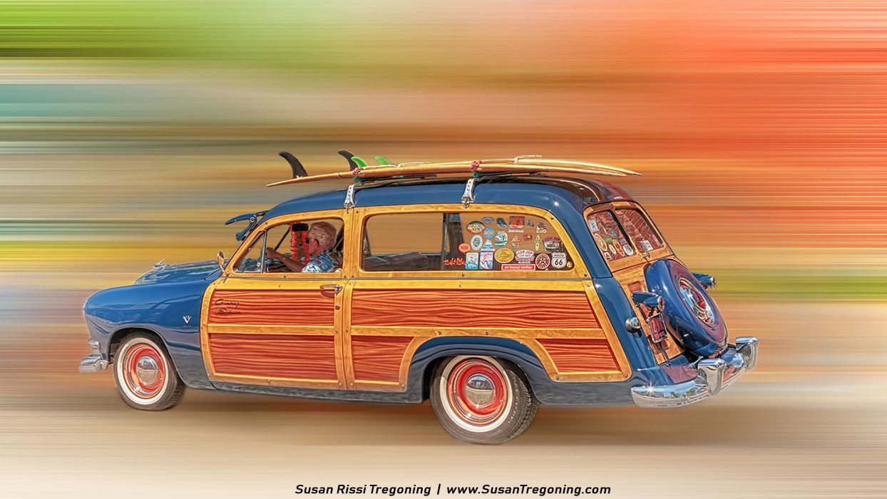 A vintage blue Ford Country Squire station wagon with wood‑panel sides drives past in a motion‑blurred, colorful background. Multiple surfboards are strapped to the roof, and the rear side windows are covered in bright travel and surf stickers. The driver wears a tropical shirt, reinforcing the beach‑culture theme.