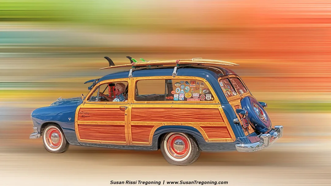 A vintage blue Ford Country Squire station wagon with wood‑panel sides drives past in a motion‑blurred, colorful background. Multiple surfboards are strapped to the roof, and the rear side windows are covered in bright travel and surf stickers. The driver wears a tropical shirt, reinforcing the beach‑culture theme. 