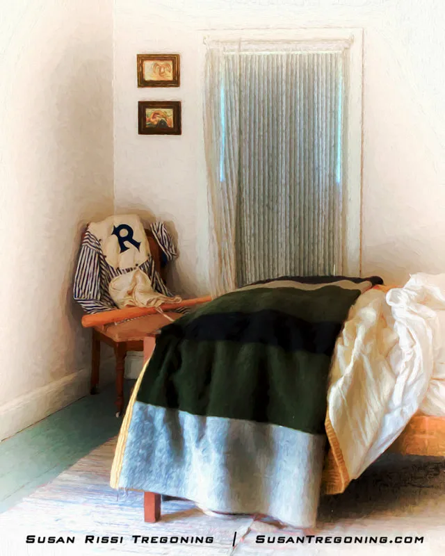 A small bedroom contains a single bed with a striped blanket, a wooden bench holding a striped shirt and a pillow with a large blue “R,” and a baseball uniform and bat placed in the corner. Soft light filters through a window with sheer curtains.
