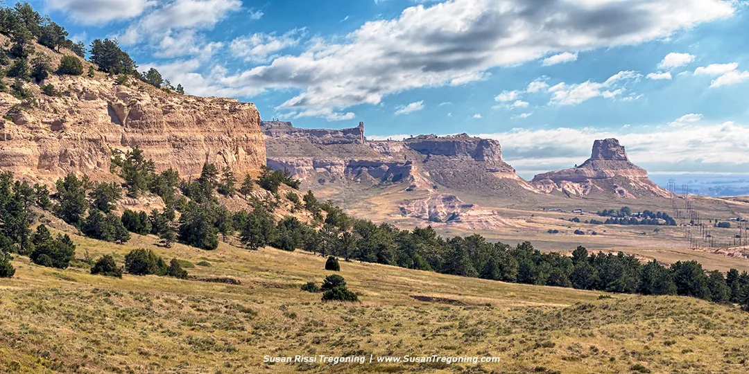 Layered cliffs and rock formations rise above grassy slopes and scattered pines, showing the backside of Dome Rock, Crown Rock, and Sentinel Rock as seen from Robidoux Pass under a partly cloudy sky.