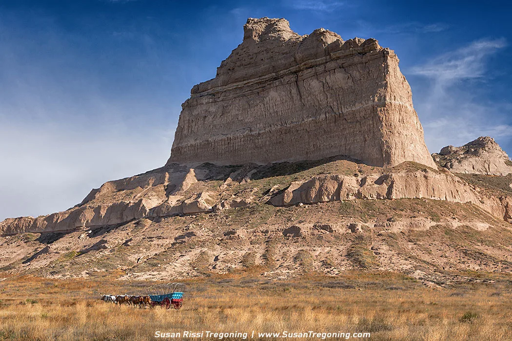 A covered pioneer wagon sits on dry grass below the towering cliffs of Scotts Bluff, with Eagle Rock rising sharply behind it under a blue sky with thin clouds.