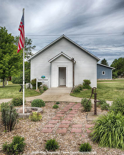 The First School of De Smet, a restored one‑room schoolhouse where early Dakota Territory students were taught, preserved today with its garden path, flagpole, and hand pump on the Ingalls Homestead grounds.