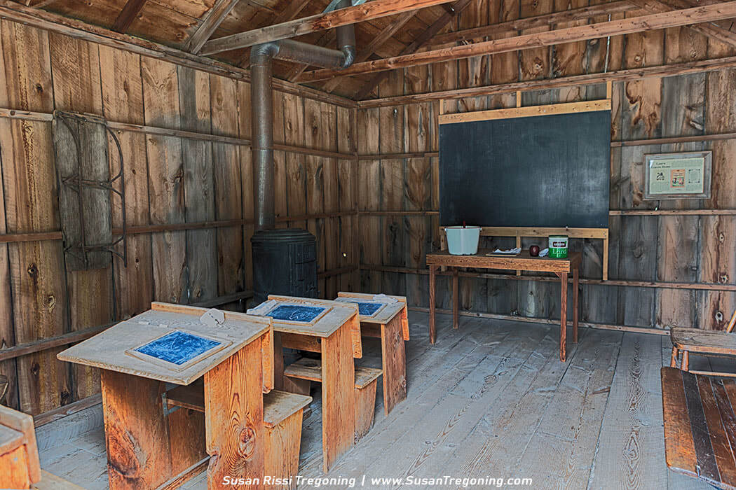The interior of the Brewster School, a rough one‑room claim shanty much like the one where Laura Ingalls Wilder taught during the winter of 1883–84, furnished with wooden desks, slate boards, a cast‑iron stove, and a simple teacher’s table.