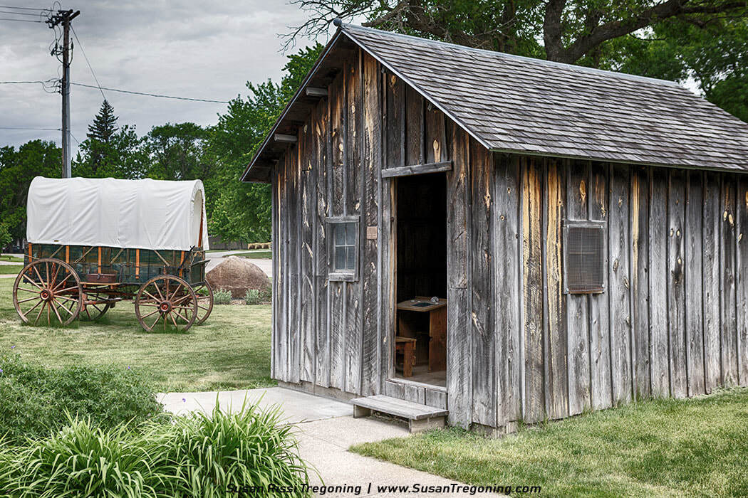 A weathered wooden claim shanty stands beside a canvas‑topped covered wagon on a grassy prairie, its open doorway revealing a simple table and chair inside, evoking the isolated Brewster School setting from Laura Ingalls Wilder’s teaching years.