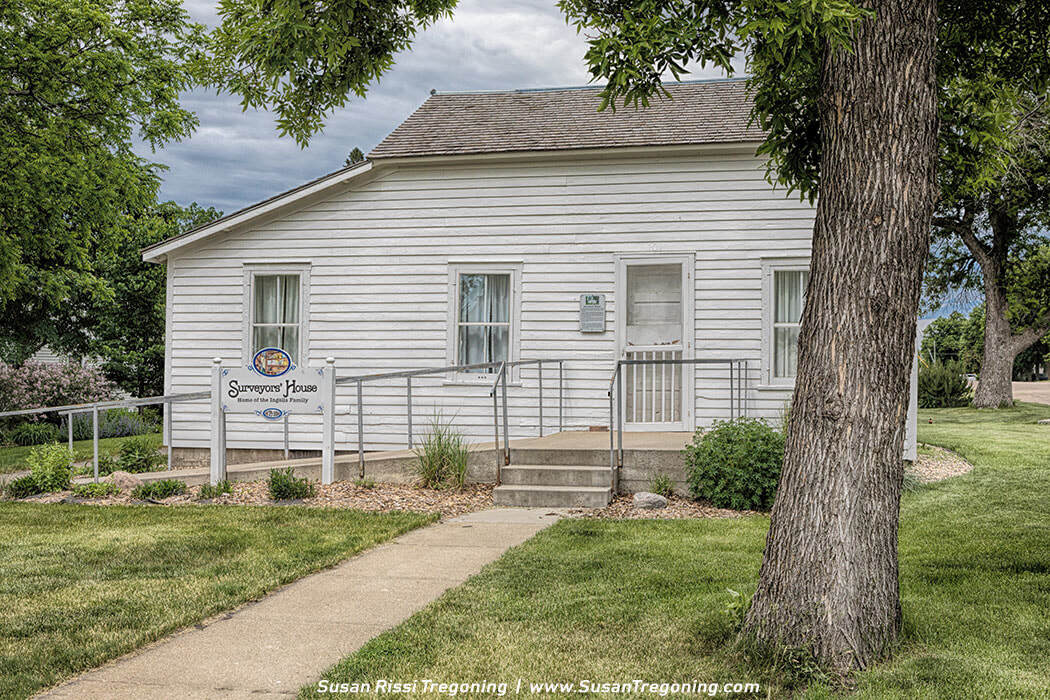 A small white wooden house known as the Surveyors’ House sits on a neatly kept lawn, with a front ramp leading to the entrance and a sign identifying it as the place where the Ingalls family lived, surrounded by leafy trees on a bright day.