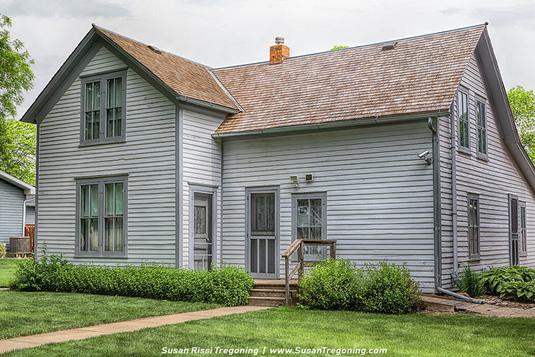 A preserved Ingalls family home in De Smet, shown as a gray wood‑sided house with a gabled roof, porch steps, and well‑kept landscaping.