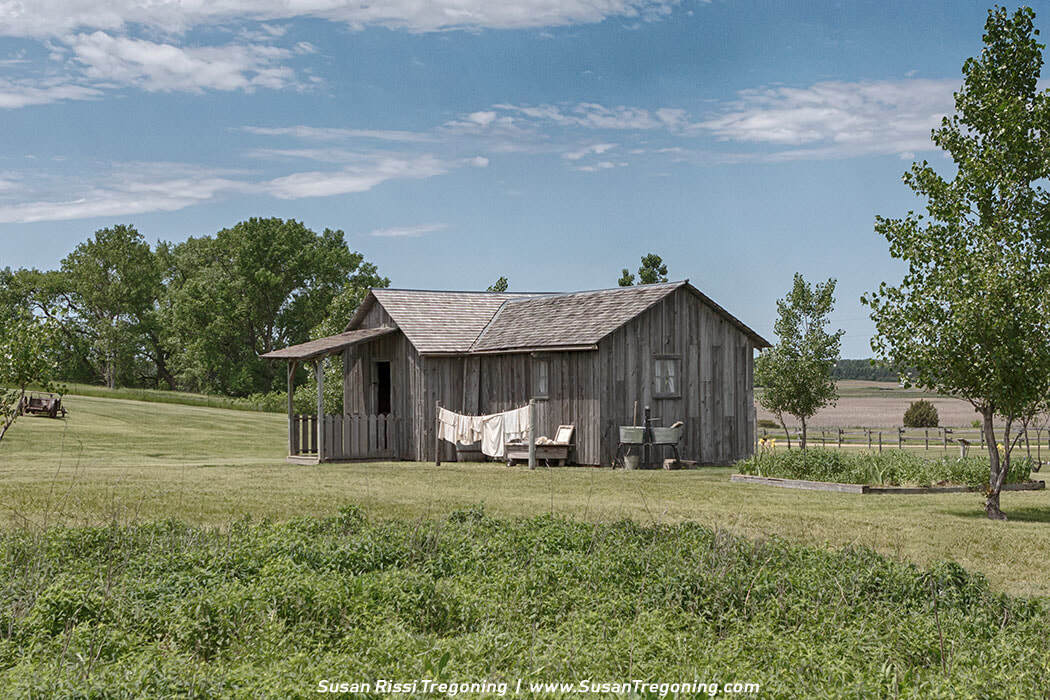 A rustic wood‑plank cabin with a shingled roof and porch, surrounded by grass, garden beds, and scattered trees, with clothes drying on a line in the yard.