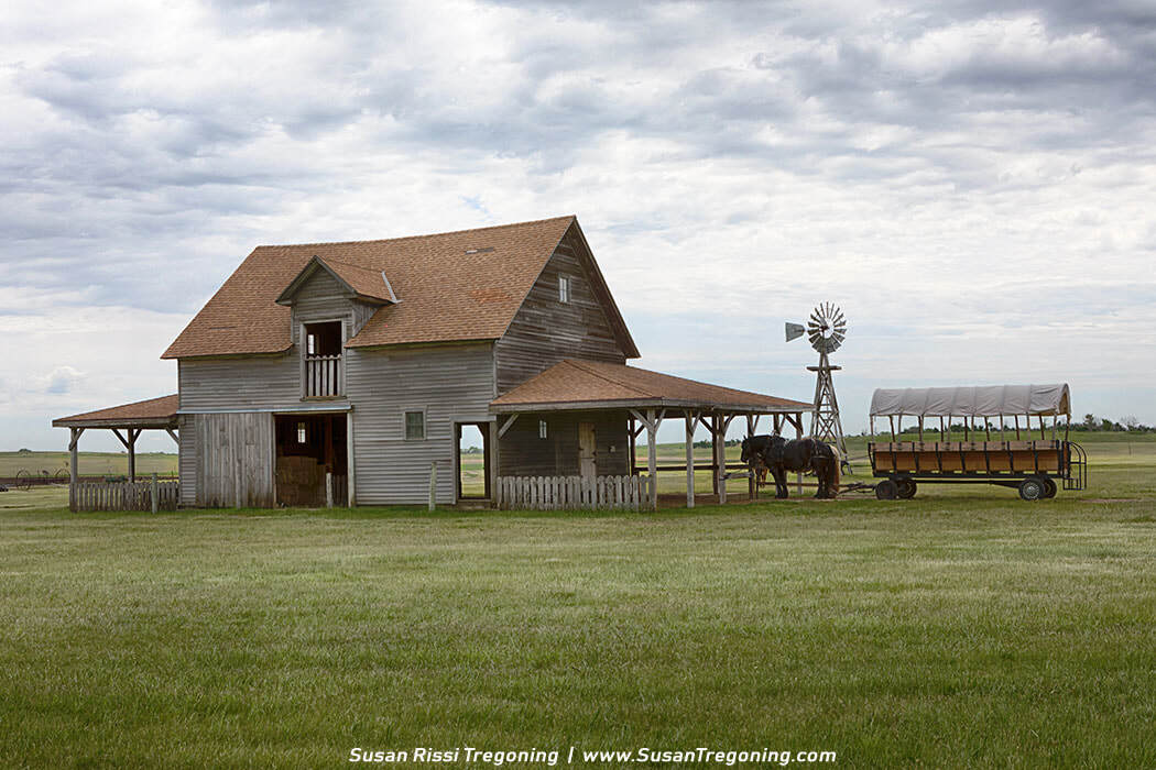 A historic barn on the Ingalls Homestead grounds, shown with its loft-level opening, surrounding grassland, a nearby windmill, and a covered wagon beneath a cloudy sky.