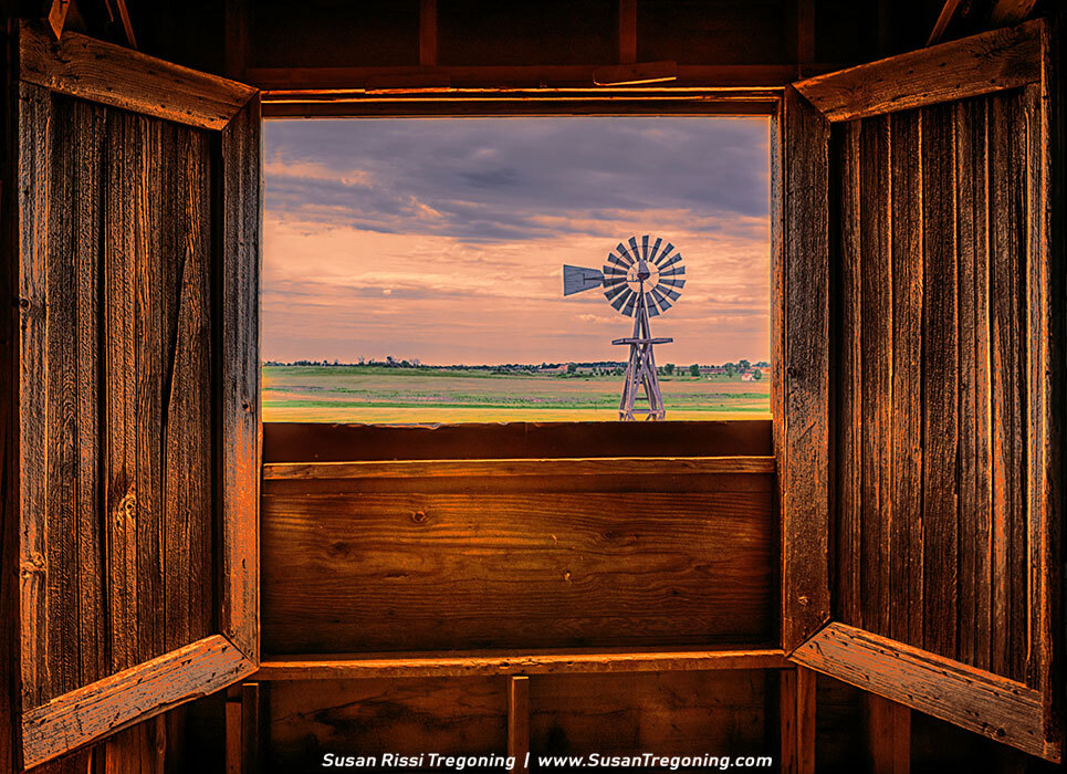 An open wooden window frame looking out onto a prairie windmill in a green field, illuminated by warm sunset light that glows across the interior boards and the partly cloudy sky.