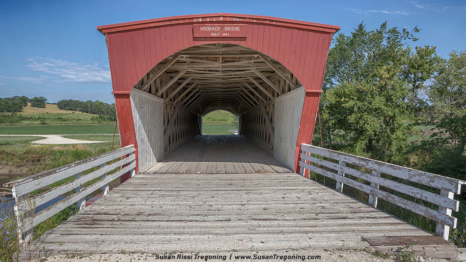 A red covered bridge labeled “Hogback Bridge, Built 1884” spans a small waterway in a rural landscape, with white interior walls, wooden plank flooring, and green fields and trees surrounding the structure.