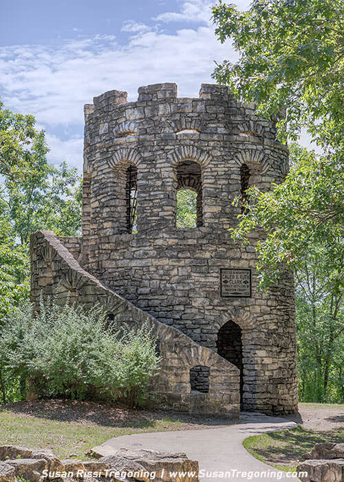A round stone tower known as Clark Tower stands among dense green trees, with arched openings, a crenellated top, and a curved exterior staircase leading to the entrance. A plaque on the wall honors Albert C. Dawson.