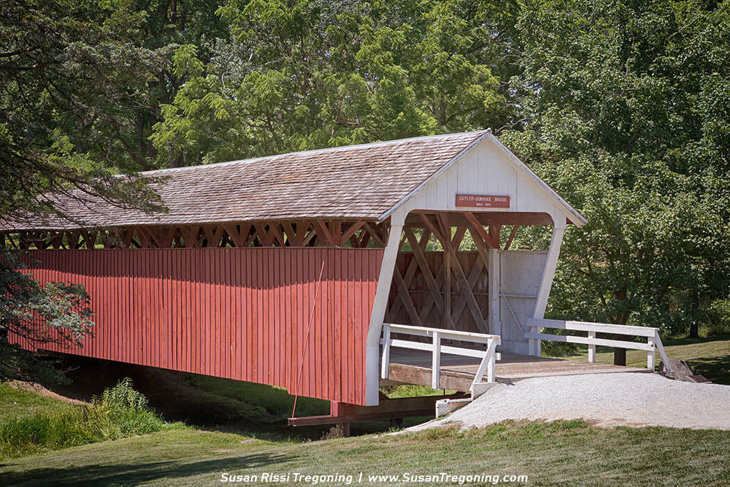 A red covered bridge labeled “Cutler-Donahoe Covered Bridge, Built 1883” spans a small stream in a lush wooded setting, with white trim, a shingled roof, and lattice trusses visible through the side openings. A gravel path leads to the entrance, surrounded by dense green trees and vegetation.