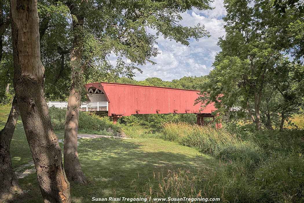 A bright red covered bridge spans a narrow creek in a lush green landscape. The wooden bridge has a pitched roof, white trim at the entrance, and sits on timber supports above the water. Trees with early‑season foliage surround the scene under a partly cloudy sky. 
