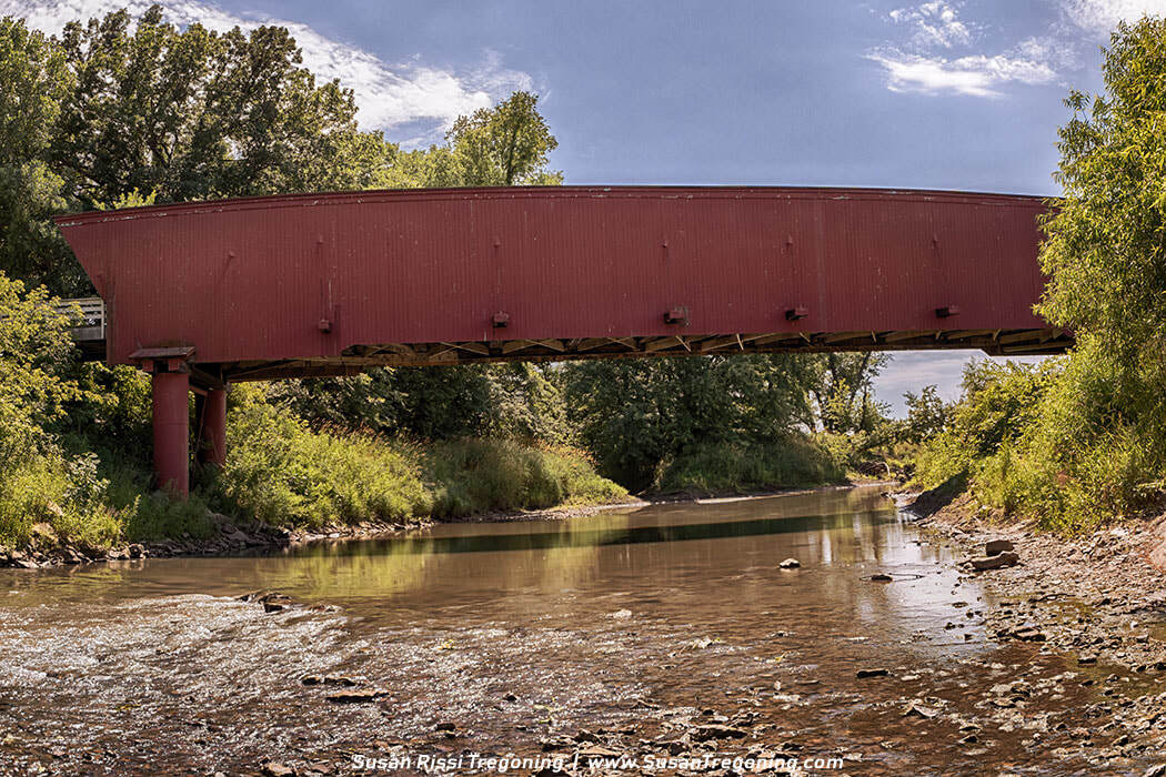 The 1880 Holliwell Covered Bridge stands in a quiet bend of the Middle River, its red siding and timber trusses anchoring the rural Iowa landscape.