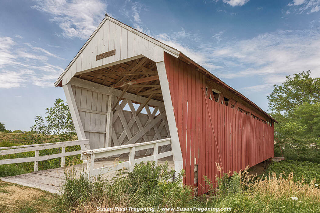 A red Imes Covered Bridge with a white entrance stands slightly elevated above a grassy slope, its wooden truss interior visible through the open portal. A short wooden ramp leads up to the bridge, and a small plaque is mounted above the entrance. Green trees and a partly cloudy sky frame the rural setting.