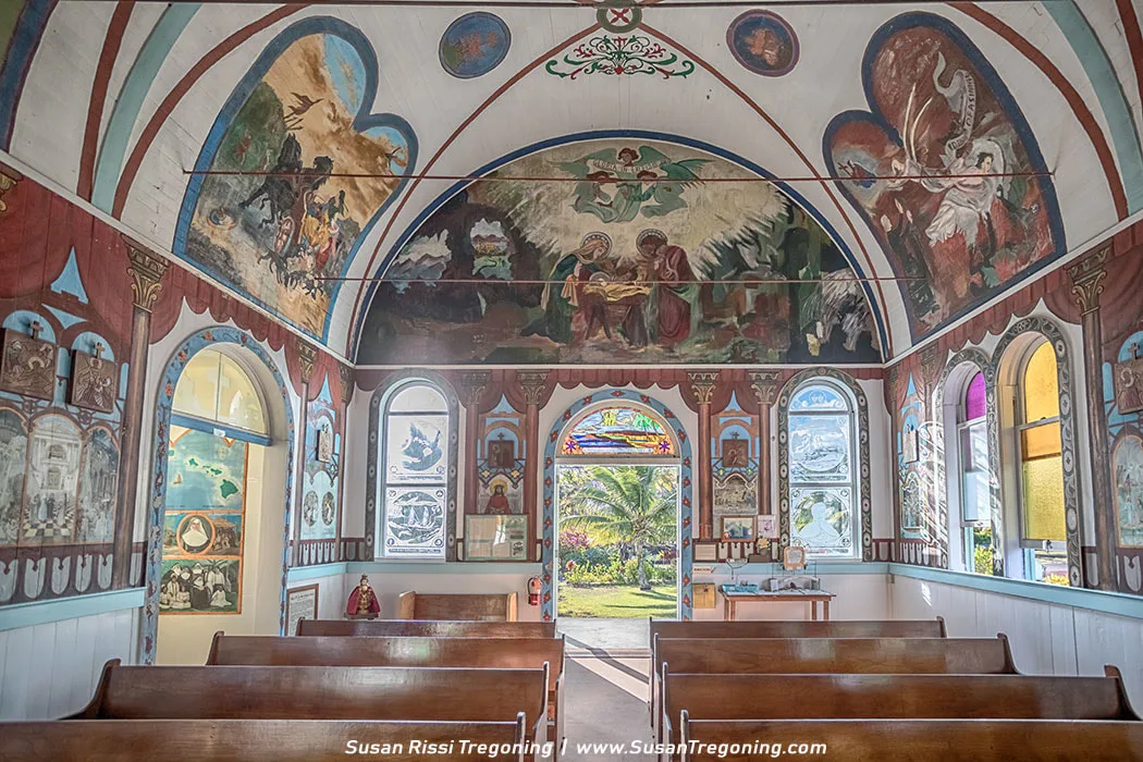 Interior of a small painted chapel with rows of wooden pews facing a mural‑covered sanctuary wall. The ceiling and upper walls are filled with colorful Catholic iconography, including angels, saints, and biblical scenes. Arched windows along the sides contain etched glass panels, and an open doorway at the back reveals bright greenery outside. The vivid murals and decorative details create an intimate, richly painted worship space.