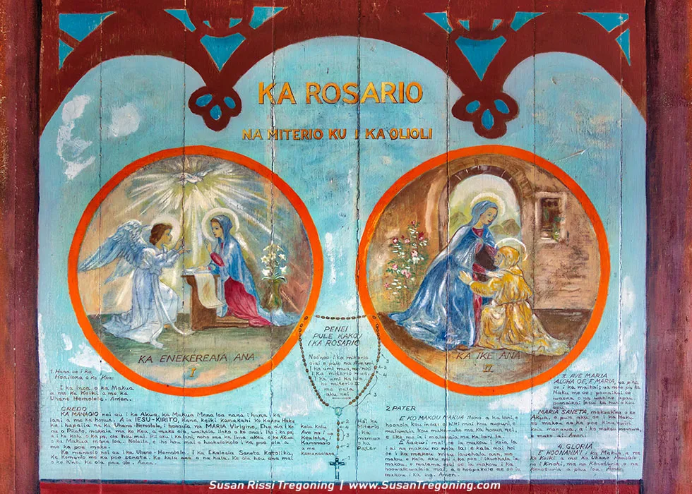 A painted mural titled “Ka Rosario” features two circular scenes from Christian tradition. The left circle shows the Annunciation, with the angel Gabriel appearing to Mary, and the right circle shows the Visitation, with Mary greeting Elizabeth. Hawaiian text surrounds the images, including prayers and instructions for reciting the rosary. Decorative borders and lettering frame the artwork, which blends Catholic iconography with Hawaiian language.