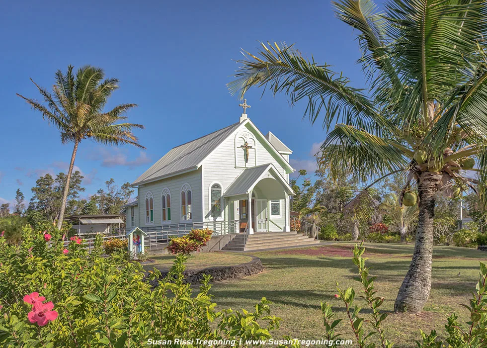 A small pale green wooden church with a steep metal roof stands among lush tropical plants and palm trees under a clear blue sky. A front porch leads to an arched entrance topped with a statue of Jesus and a cross, and arched windows line the sides of the building. Bright greenery and flowering plants surround the church, creating a peaceful tropical setting.