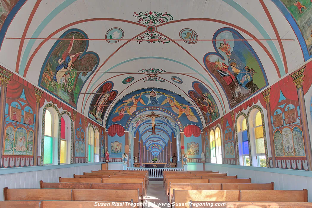 Interior of Star of the Sea Painted Church with rows of wooden pews facing an altar that holds religious statues. A painted trompe l’oeil backdrop in soft blue tones rises behind the altar, while the ceiling above is covered with Catholic iconography and decorative motifs. Colorful murals and faux‑architectural details line the walls, and arched windows along the sides bring natural light into the small sanctuary.