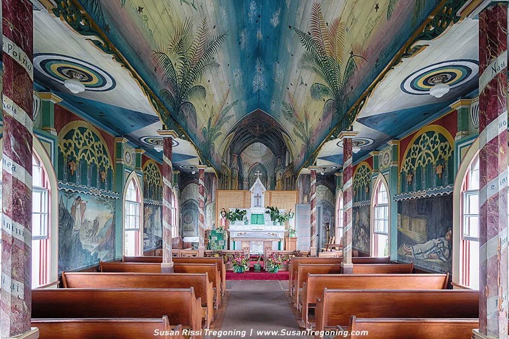 Interior of St. Benedict Painted Church with wooden pews facing a small altar decorated with green cloths and flowers. The ceiling is painted with a sky scene of blue tones, palm fronds, and stars, while the walls feature colorful religious murals and faux‑marble arches. Natural light enters through arched windows, illuminating the vivid artwork covering nearly every surface.