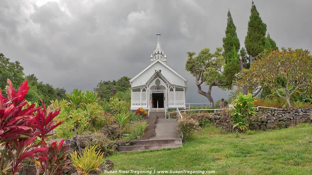 A small white historic church with a pointed steeple and cross stands at the end of a straight paved walkway, surrounded by lush tropical plants and a low stone wall. Dark, dramatic clouds fill the sky behind the building, and the year “1853” appears above the entrance. This is St. Benedict’s Painted Church in South Kona, Hawaiʻi.