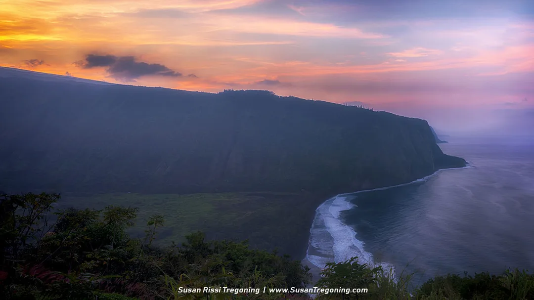 A steep, vegetation‑covered cliff drops toward the ocean as waves roll in below. The sky glows with warm sunset colors—orange, pink, and purple—and soft haze surrounds the cliff, creating a calm, scenic coastal view.