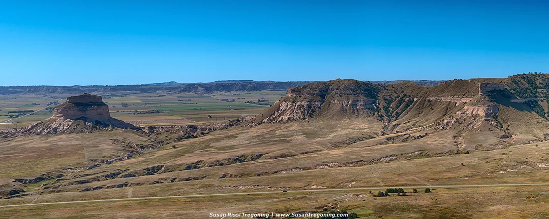 A wide panoramic view of Scotts Bluff shows Dome Rock rising from the plains, with layered cliffs, open farmland, and distant hills beneath a clear blue sky.