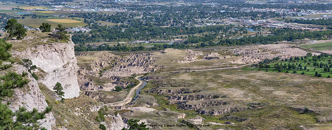 A high vantage point atop Scotts Bluff overlooks a winding dirt road, a narrow waterway, and the town and farmland stretching across the valley below.