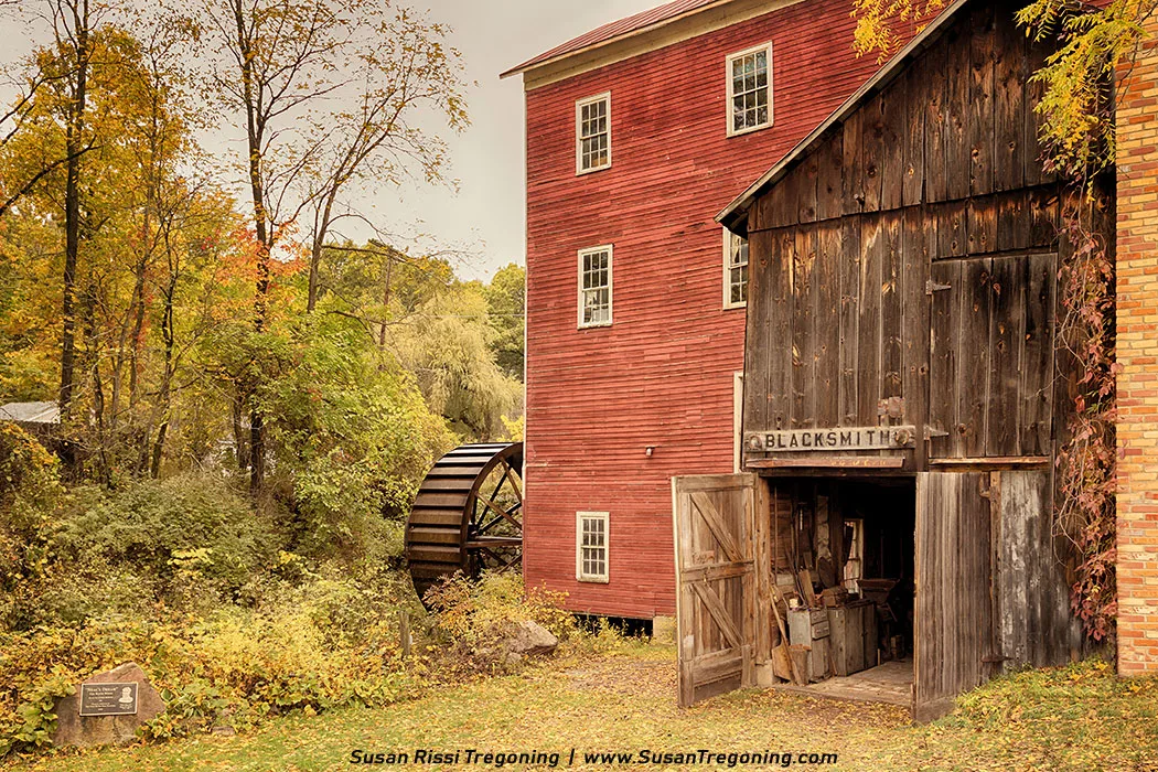 A red historic mill with a large wooden water wheel stands beside a rustic blacksmith shop at Bowens Mills. The blacksmith building has open doors showing tools inside, and both structures are surrounded by autumn trees. A small plaque sits on a rock in the foreground, marking the site’s historical significance.