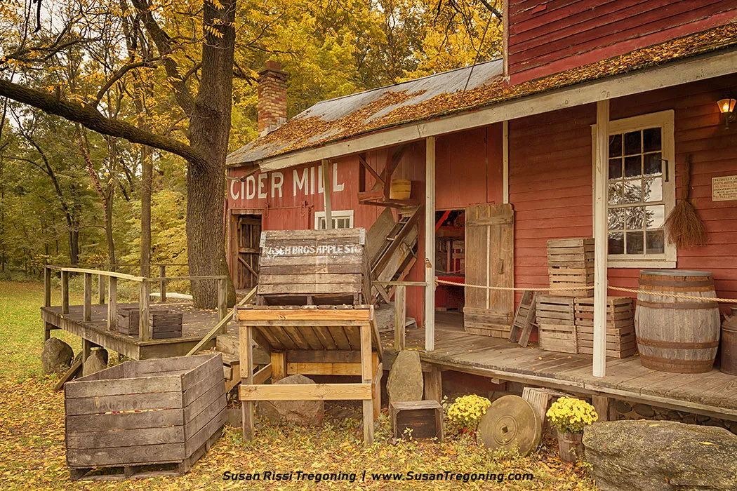 A red cider mill with white trim stands among vibrant autumn trees. Wooden crates, barrels, and apple‑processing equipment sit on the porch and in the yard, adding to the rustic harvest setting. The scene is surrounded by warm fall foliage, creating a nostalgic rural atmosphere.