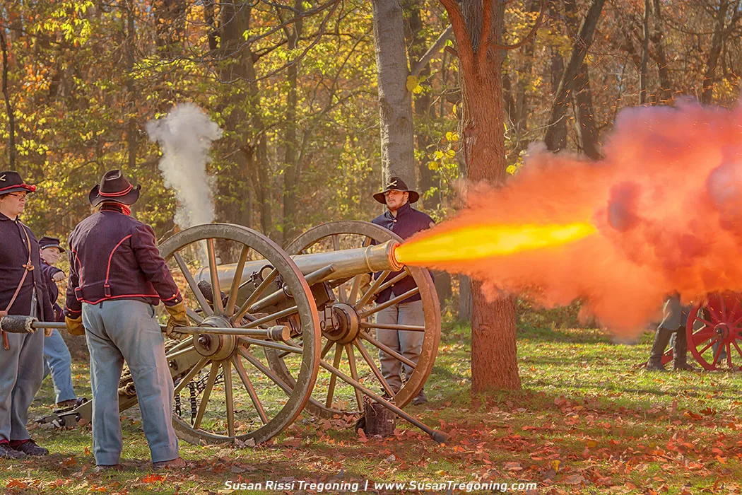 Union soldiers in Civil War uniforms fire a cannon during a reenactment at Bowens Mills in Yankee Springs, Michigan. A bright burst of flame and smoke erupts from the cannon as reenactors stand positioned around it in an autumn woodland setting.