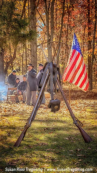Three vintage rifles are arranged as a tripod in the foreground, with leather straps and a satchel hanging from them. Behind the rifles, Civil War reenactors in period uniforms gather in an autumn woodland, with sunlight filtering through the trees and a historic American flag displayed to the right.
