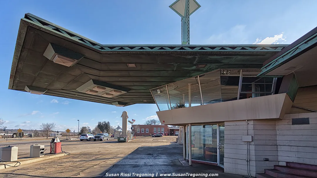A vintage gas station with a dramatic, futuristic overhang resembling a wing extends over a glass‑walled office area. Gas pumps sit in the foreground, and the bold, angular design reflects mid‑century Googie architecture. Vehicles and small buildings appear in the background beneath a clear sky.