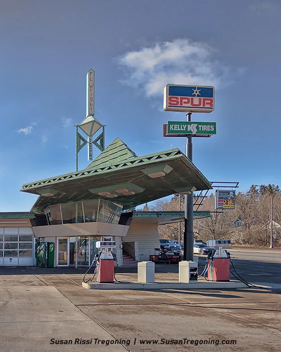 A uniquely designed gas station features a bold green angular roof and a central tower labeled “FLWright.” Two gas pumps stand in front of the building, which displays Spur and Kelly Tires signage.