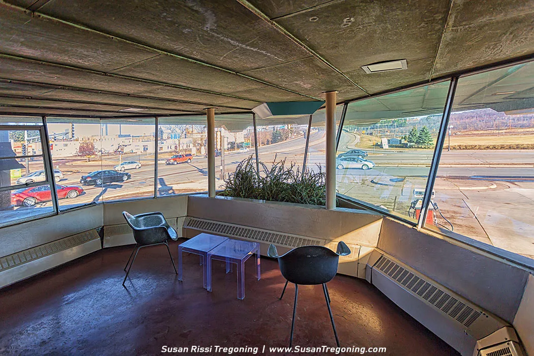 The interior of Wright’s Waiting Station features large slanted glass windows overlooking a busy street. Two modern black chairs and a clear coffee table sit on a reddish‑brown floor beside a planter of tall grasses. The geometric ceiling has recessed lighting, and built‑in radiators line the walls. Vehicles and buildings are visible outside through the expansive windows.