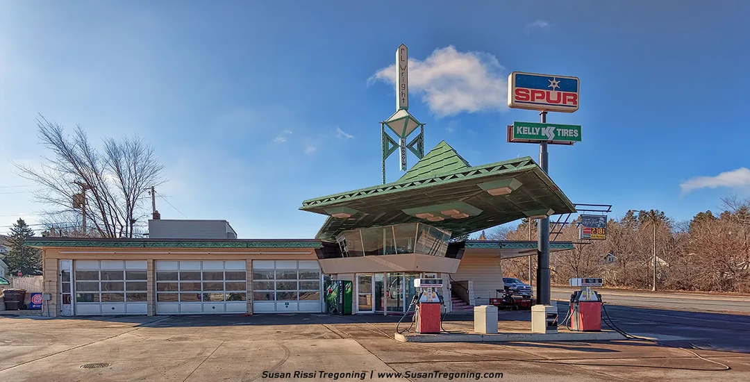 A vintage-style gas station with Frank Lloyd Wright–inspired architecture features a green angular canopy topped with a tall spire labeled “F.L. Wright.” Two gas pumps stand in front of the building, which includes several garage doors and signage for Spur and Kelly Tires. 