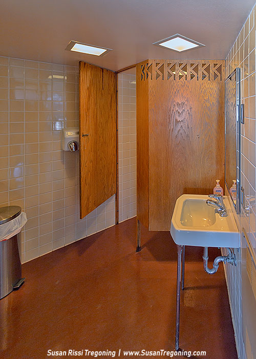 A mid‑century modern restroom interior features a single white porcelain sink with exposed plumbing, two faucets, and a mirror above it. Beige square tiles cover the walls, and a reddish‑brown floor leads to a wooden stall door with a geometric cut‑out panel. A hand dryer is mounted beside the stall, and a stainless steel trash can sits near the entrance. Recessed square ceiling lights illuminate the space.