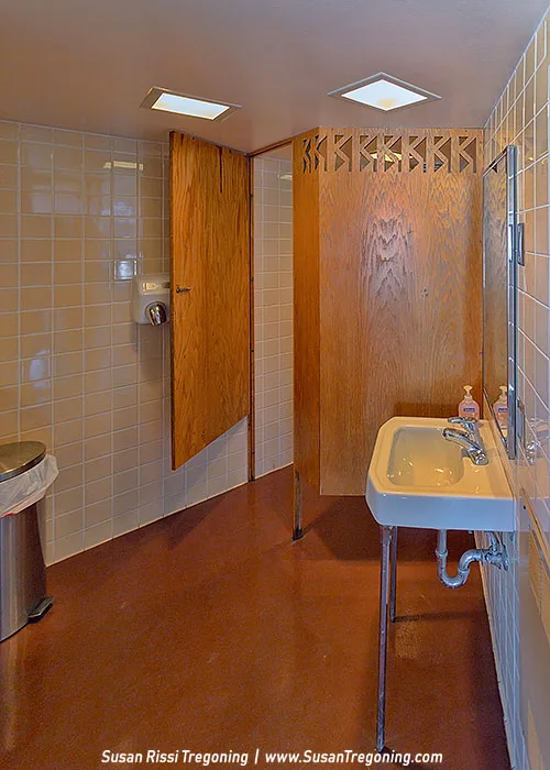A mid‑century modern restroom interior features a single white porcelain sink with exposed plumbing, two faucets, and a mirror above it. Beige square tiles cover the walls, and a reddish‑brown floor leads to a wooden stall door with a geometric cut‑out panel. A hand dryer is mounted beside the stall, and a stainless steel trash can sits near the entrance. Recessed square ceiling lights illuminate the space.