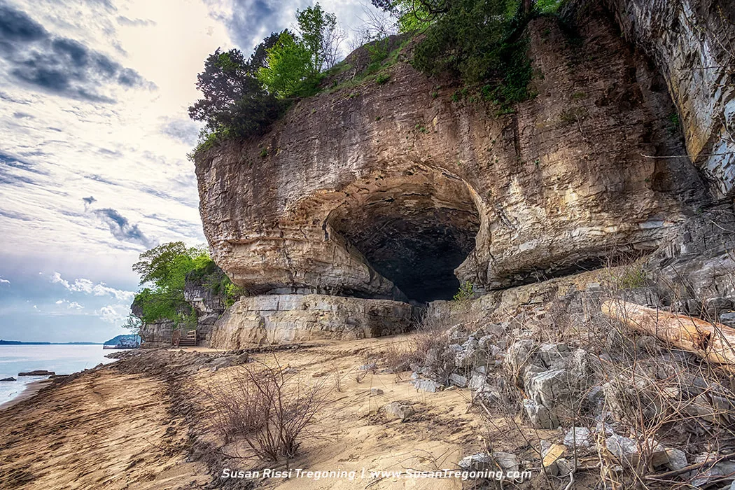A large natural cave opens in a tall limestone cliff along a sandy shoreline. Layered rock formations rise above the cave, topped with green vegetation and trees. Sunlight filters through a partly cloudy sky, illuminating the rock face and the quiet water beside the shore.