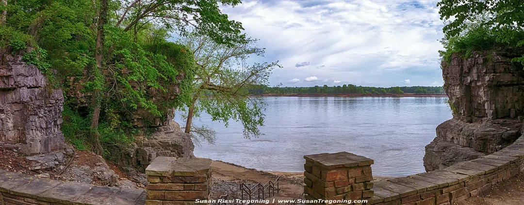 A panoramic view of the Ohio River is seen from a stone overlook at the top of a stairway leading down toward a cave. Rocky cliffs and dense green vegetation frame the river as it stretches into the distance beneath a partly cloudy sky.
