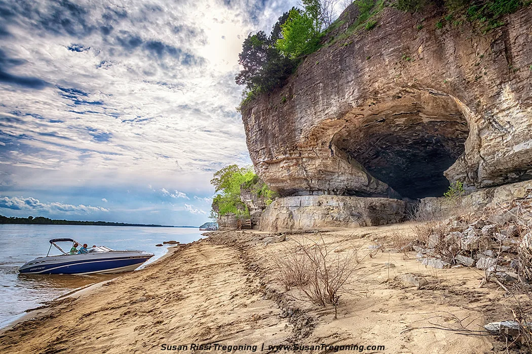 A tall limestone bluff with a large natural cave at its base rises beside the Ohio River. A sandy shoreline stretches across the foreground, where a small motorboat is pulled up on the sand. Vegetation grows along the top and edges of the rock formation, and sunlight filters through a partly cloudy sky, reflecting off the calm water.