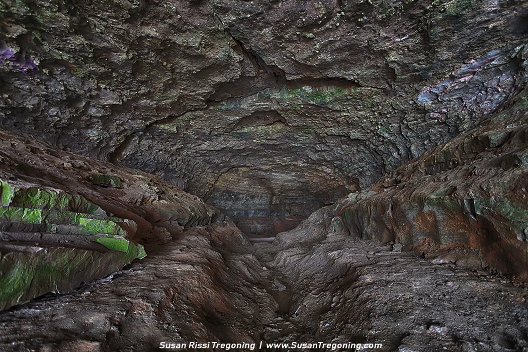 A narrow cave passage with a rough, layered rock ceiling and walls shows deep grooves carved into the uneven floor. Shades of brown, gray, and green highlight the textured stone, with patches of moss adding color as light reveals the depth and contours of the formation.