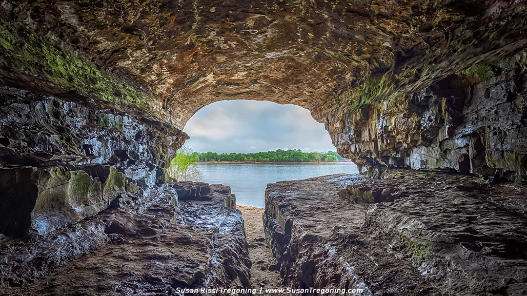 View from inside a rocky cave looking out toward a calm river, with layered stone walls and a grooved floor in shadow. Sunlight brightens the tree‑lined shoreline and partly cloudy sky beyond the cave’s wide opening.