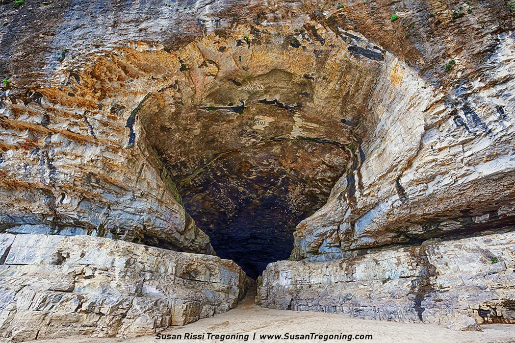 A wide natural cave entrance opens in a tall limestone cliff, with layered sedimentary rock walls and a sandy floor at the base. Sunlight highlights the tan and gray rock textures while trees and vegetation grow along the top of the formation beneath a partly cloudy sky.