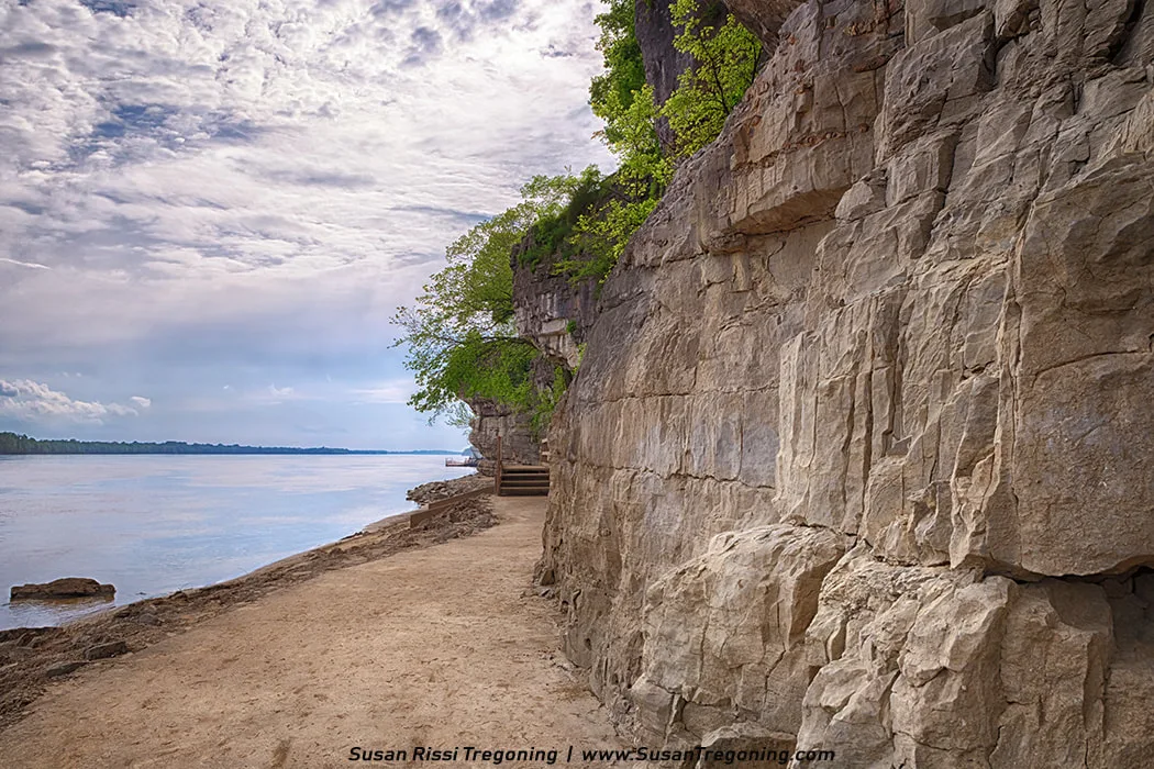 A rocky cliff of layered sedimentary stone rises beside a calm river, with a dirt path running along its base. Green foliage grows from the top and sides of the cliff, and the river stretches into the distance beneath a partly cloudy sky.