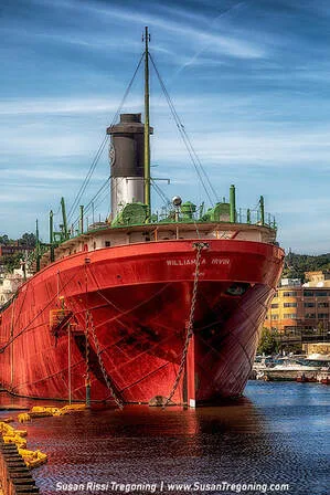 The 610‑foot Great Lakes freighter SS William A. Irvin is docked along the waterfront, with its long hull and superstructure visible beneath cloudy skies. The surrounding area includes industrial and museum‑ship facilities.